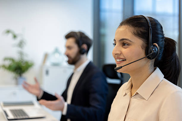 A smiling woman wearing a headset works at a call center alongside a colleague, both engaged in a discussion.